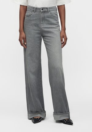 Persona che indossa jeans grigi a gamba larga con risvolti, top bianco testurizzato e scarpe nere a punta con borchie color argento, in piedi contro uno sfondo semplice.