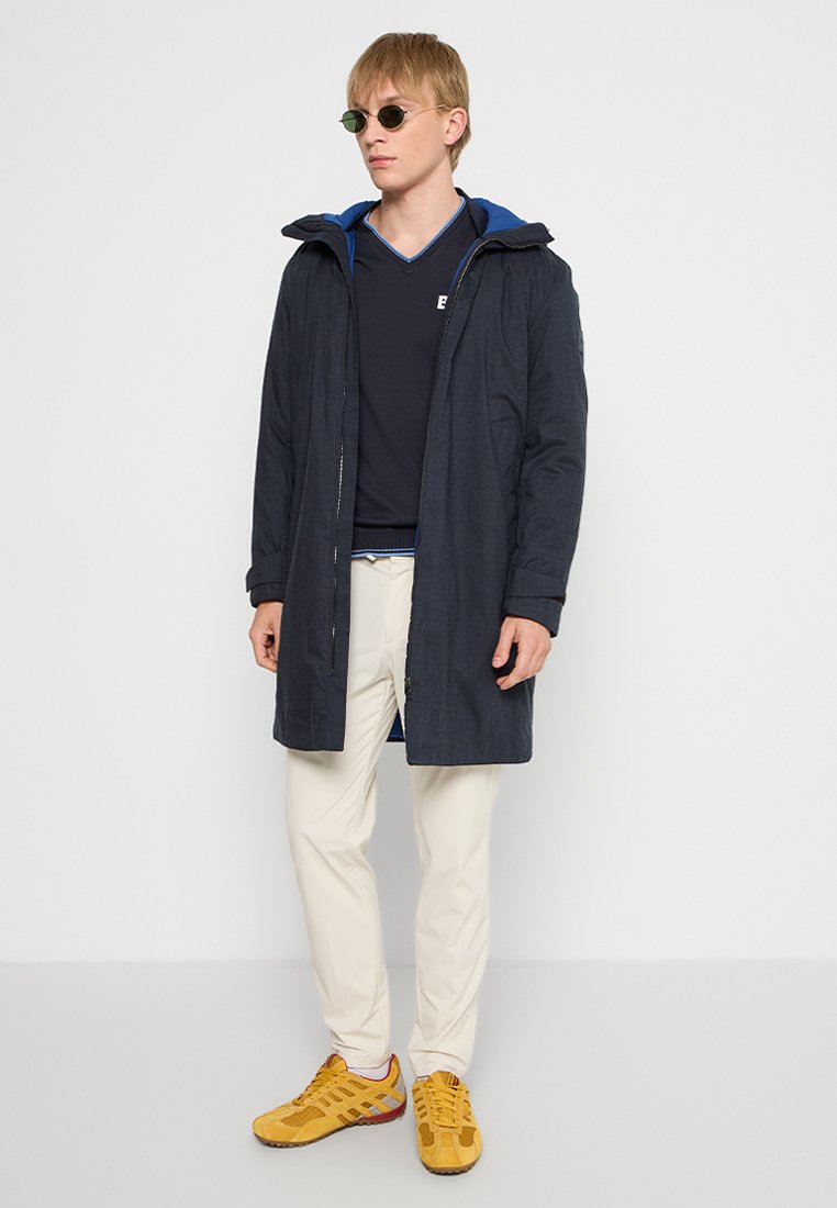 Manteau long marine avec une capuche, doté d'une doublure intérieure bleue. Porté sur une chemise noire et un pantalon crème, associé à des baskets jaunes.
