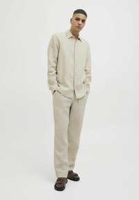 Camisa de lino beige claro con cuello, frente de botones y corte relajado, combinada con pantalones a juego y sandalias oscuras. Textura de tela suave.