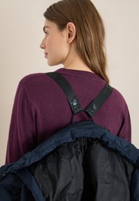 Kvinde med langt brunt hår iført en lilla sweater og en marineblå jakke, der hænger over hendes skuldre med sorte stropper.