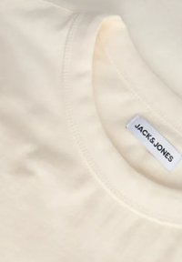 Lys beige t-shirt med rund hals, med et label der siger "JACK & JONES." Glat tekstur og minimalistisk design.