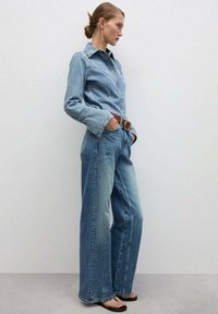 Jeans srajca s ostrim ovratnikom, gumbom za zapiranje, dolgimi rokavi; v kombinaciji z širokimi modrimi kavbojkami in rjavim pasom, oblečenimi s črnimi sanalkami.