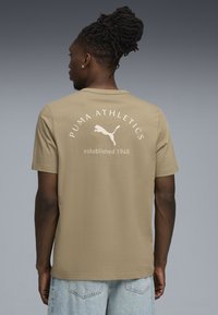 Beżowa bawełniana koszulka z krótkim rękawem, z napisem "PUMA ATHLETICS" i logo w białym kolorze na plecach. Luźny krój i minimalistyczny design.