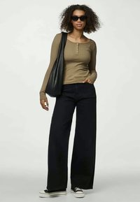 Braunes, geripptes Langarmoberteil, schwarze, weit geschnittene Hose, schwarze Sonnenbrille und schwarze Einkaufstasche, dazu schwarze und weiße Sneaker.