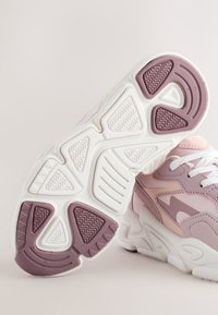 Chaussures de sport roses et blanches avec semelles épaisses, motifs de bande de roulement détaillés et lacets blancs sur un fond clair et neutre.