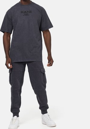 Hombre vistiendo una camiseta Kani oversized en gris oscuro, pantalones jogger cargo a juego y zapatillas blancas, de pie contra un fondo blanco.