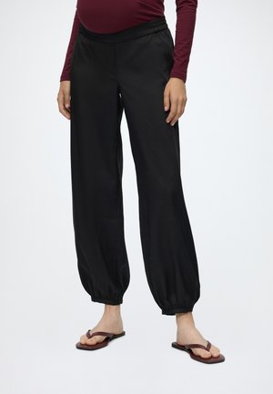 Schwangere Person trägt schwarze Hosen mit elastischem Saum, ein bordeauxfarbenes Langarmshirt und bordeauxfarbene Flip-Flops, steht vor weißem Hintergrund.