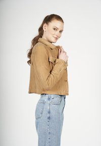 Tan suede cropped jack met een kraag, een voorzak en ritsdetails, gecombineerd met lichtblauwe high-waisted jeans.