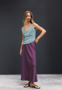 Gaiši zils tanktops kopā ar garu sarkanu un tumši zilu svītrainu svārku, kas ir brīva piegriezuma. Modelis ir basām kājām, stāvot pret tumšu fonu.