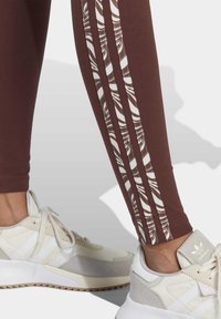Bruna leggings med vita mönstrade sidostråk, kombinerade med vita sneakers med ljusgrå accenter och en texturerad sula.