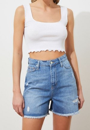 Vrouw draagt een wit cropped tanktop en hooggetailleerde blauwe denim shorts met rafelige zomen en distressed details.