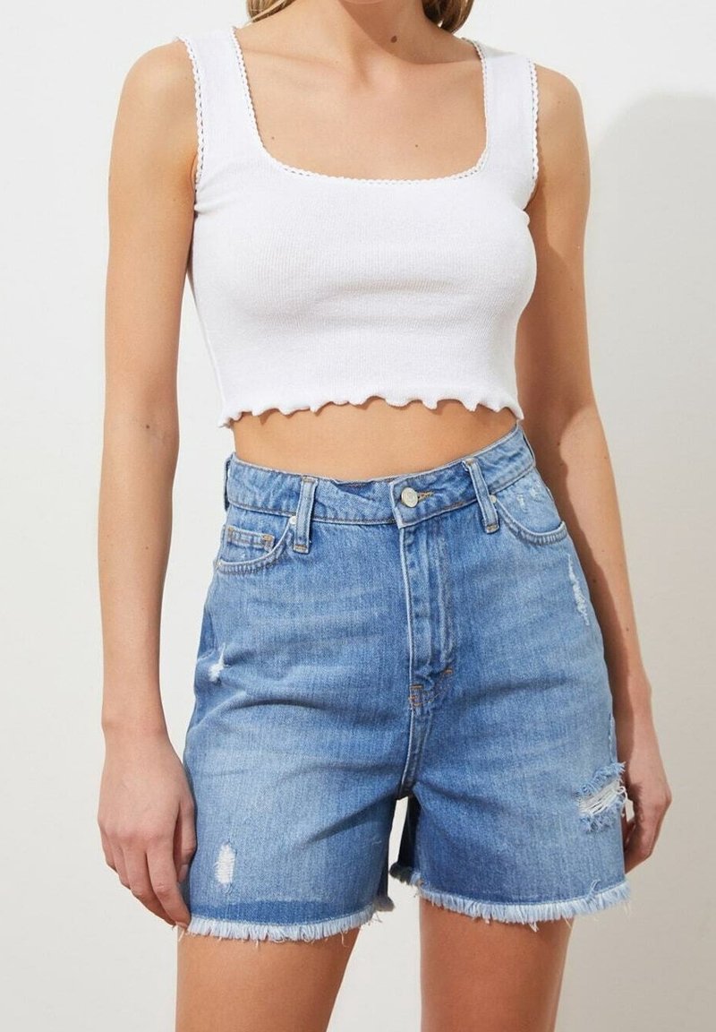 Femme portant un débardeur blanc court et un short en denim bleu taille haute avec des bords effilochés et des détails usés.