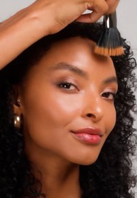 Femme aux cheveux bouclés applique du fond de teint sur son front à l'aide d'un grand pinceau, portant des boucles d'oreilles dorées et un maquillage des yeux subtil.