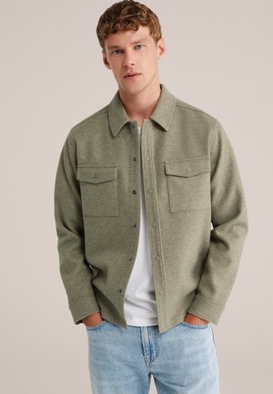 Zelená košile-jacket z vlněné směsi s designem na knoflíky, dvěma náprsními kapsami a špičatým límcem. Nášita přes bílou košili a světle modré džíny.