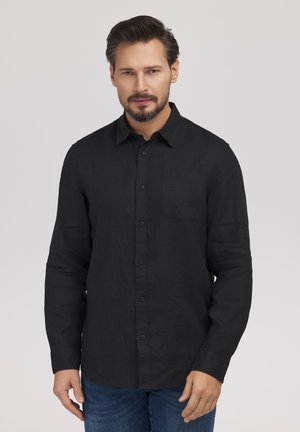 Mężczyzna o krótkich ciemnych włosach i brodzie, ubrany w czarną, zapinaną na guziki koszulę z długim rękawem i niebieskie dżinsy, stojący na tle jednolitego jasnego tła.