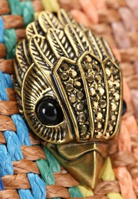 Brązowy brosza w kształcie ryby ozdobiona czarnymi kamieniami i misternym detalem piór, na tle wielokolorowego tkankowego tła.