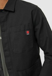 Svart knapp-up skjorta med en bröstficka och en röd cirkellogo, tillverkad av slitstark tyg med en slät yta och en klassisk passform.