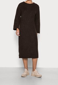 Braunes Strickkleid mit rundem Ausschnitt und langen Ärmeln, kombiniert mit hellbeigen Sneakern. Das Kleid hat eine lockere Passform und eine Länge bis zur Mitte der Wade.