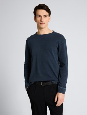 Jeune homme aux cheveux bruns courts, portant une chemise bleu foncé à manches longues et un pantalon noir, debout avec une main dans la poche.