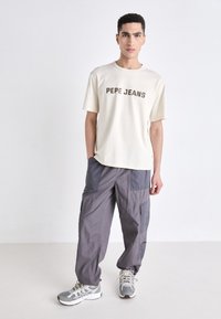 Kremfarget t-skjorte med korte ermer som har "PEPE JEANS" trykket i brunt. Grå cargo-bukser med flere lommer. Grå og hvite sneakers.