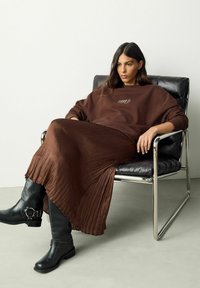 Brauner übergroßer Pullover mit Schriftzug, plissierter brauner Rock und schwarze kniehohe Stiefel. Sitzend auf einem schwarzen Stuhl mit verchromten Beinen, neutraler Hintergrund.