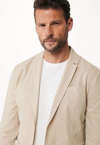 Beige leichte Jacke mit Notchkragen, einer Tasche und weicher Textur. Über einem schlichten weißen T-Shirt getragen, das eine strukturierte Passform aufweist.