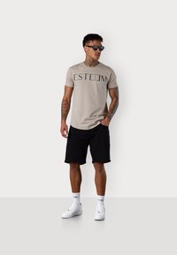 T-shirt beige à manches courtes avec inscription noire "ESTEEM", associé à un short noir et des baskets blanches. Modèle portant des lunettes de soleil noires.