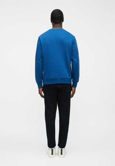 Άνδρας γυρισμένος με την πλάτη, φοράει μπλε φούτερ, μαύρο παντελόνι και λευκά αθλητικά με μαύρη και πράσινη λωρίδα, σε απλό φόντο.