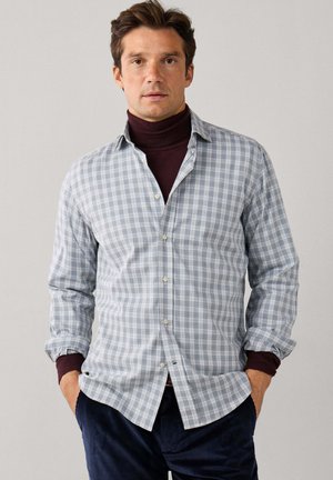 Man wearing light gray checkered shirt over dark burgundy turtleneck with hands in pockets against plain background.