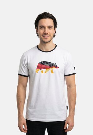 Mann mit mittellangem, dunklem Haar lächelt, trägt ein weißes T-Shirt mit einem geometrischen Bären in Schwarz, Rot und Gelb. Schlichter, heller Hintergrund.
