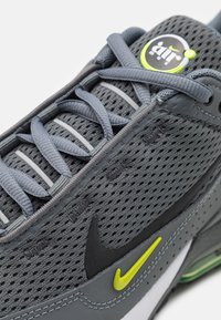 Zapatilla deportiva gris con parte superior de malla, diseño curvado, acentos en verde lima, símbolo de Nike en negro y sistema de cordones en capas.