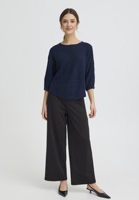 Haut en maille bleu marine avec manches larges trois-quarts, associé à un pantalon noir à jambes larges et des talons pointus noirs, sur un fond clair.