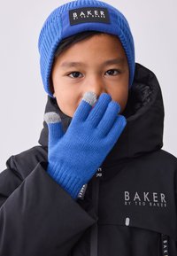 Kék kötött kesztyű szürke ujjvégekkel, egy gyermek viseli, fekete márkás dzsekivel és texturált kötött kék sapkával párosítva.