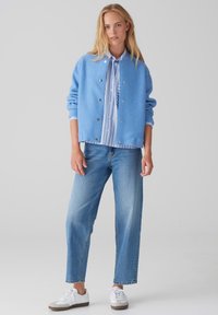 Giacca azzurro chiaro, texturizzata, con bottoni argentati sopra una camicia a righe azzurro chiaro e bianca, abbinata a jeans in denim azzurro chiaro e sneaker bianche.