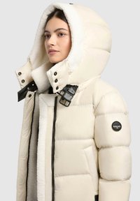 Puffer jacket in cream with a shearling-lined hood. Features a black strap with buckle and a logo patch on the left sleeve. Zipper closure.