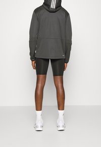 Sudadera con cremallera gris oscuro con tres rayas blancas en la capucha, combinada con pantalones cortos de ciclismo negros y zapatillas deportivas blancas.