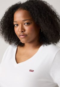 Balta V-veida kakla T-krekls no kokvilnas ar mazu sarkanu Levi's logotipa uzšuvumu uz krūtīm. Īsas piedurknes, mīksta tekstūra.