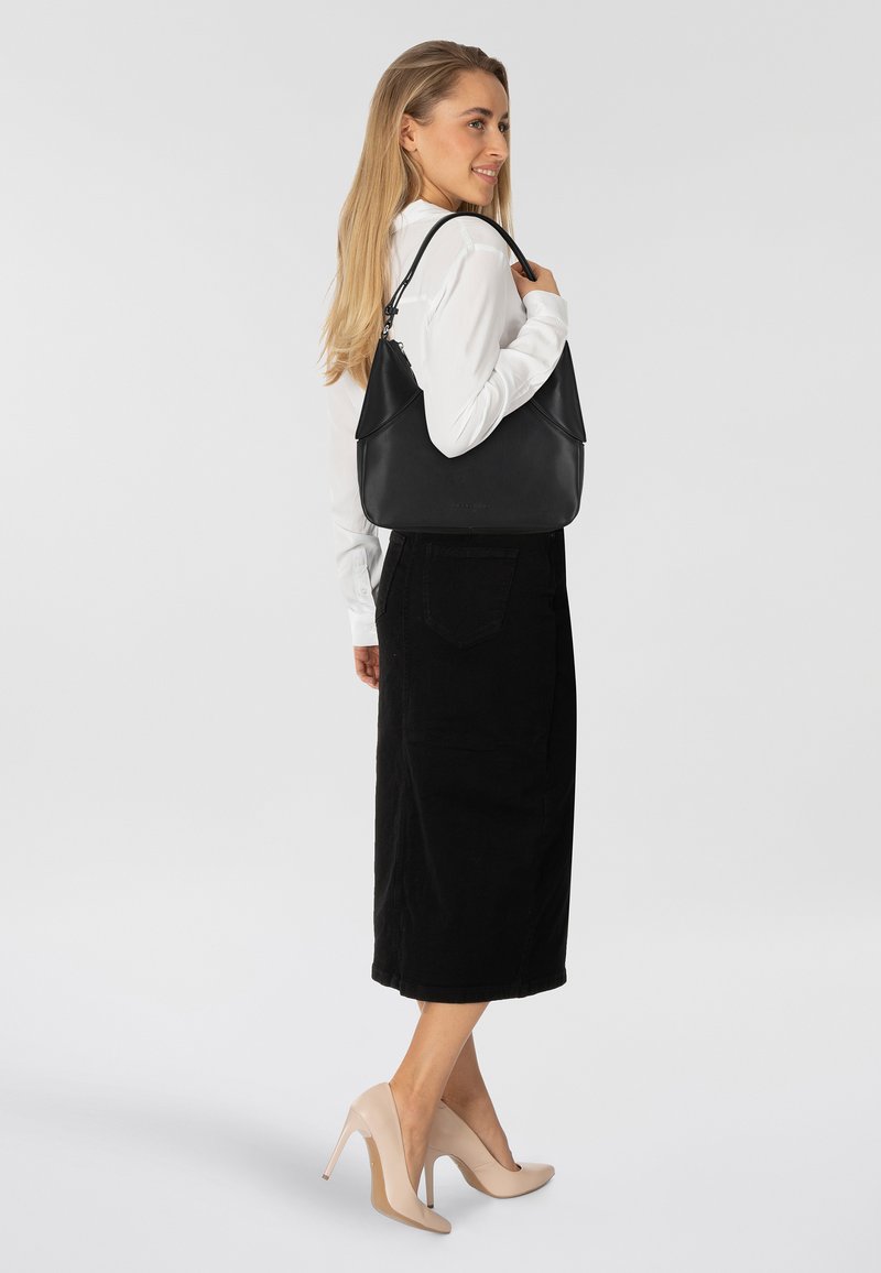 Borsa a tracolla in pelle nera con una forma morbida e parte superiore ripiegata. Indossata sulla spalla, abbinata a una camicia bianca e una gonna nera.