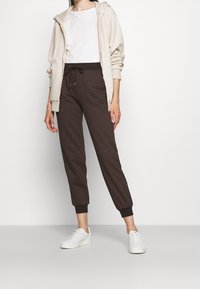 Kvinna klädd i beige zip-up hoodie, vit t-shirt, mörkbruna mönstrade joggers med dragsko och vita sneakers som står framför en enfärgad bakgrund.