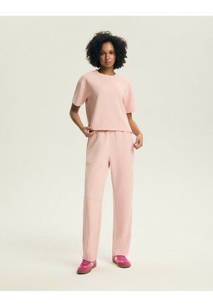 Kobieta stojąca w jasnoróżowej, dopasowanej koszulce i spodniach oraz w jasnoróżowych sportowych butach na jednolitym tle.