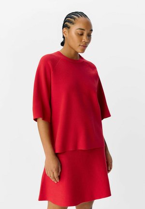 Top rosso in maglia con maniche larghe, scollo a girocollo e texture morbida, abbinato a una gonna rossa coordinata con finitura liscia.