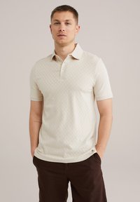 Beige polo shirt with a textured pattern, buttoned collar, short sleeves, and a small logo at the hem; paired with dark brown shorts.