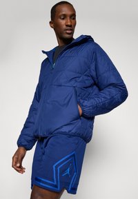 Mornarsko modra prešitna jakna z zadrgo spredaj in kapuco. Ima elastične manšete in sprednji žep. Nosijo jo z usklajenimi mornarsko modrimi kratkimi hlačami z modrimi poudarki.