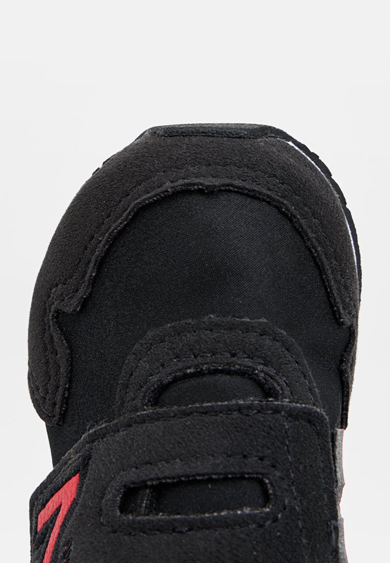 Chaussure de sport noire avec une tige en tissu, sangle Velcro, bout renforcé et accents rouges. Surface texturée pour une adhérence améliorée.