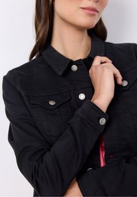 Chaqueta de denim negro con un diseño clásico, botones a presión, bolsillos en el pecho y un acabado ligeramente texturizado. Los acentos incluyen un forro rojo en contraste.