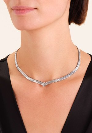 Collier en argent avec un motif en arête de poisson, doté d'un fermoir délicat. Texture lisse, porté près du cou contre un haut sombre.