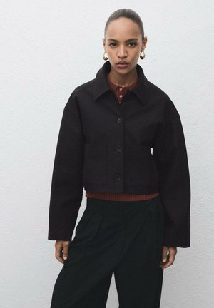 Cropped black jacket with a collar, front button closure, two pockets, and loose sleeves, layered over a burgundy top and dark green pants.