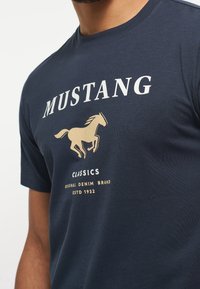 Námořnická modrá bavlněná tričko s krémovým a zlatým grafichem koně a textem "MUSTANG CLASSICS" a "ORIGINAL DENIM BRAND."