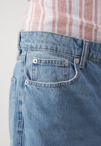 Gros plan sur une personne portant un jean en denim bleu clair et une chemise rayée blanche et rose rentrée dans la taille.