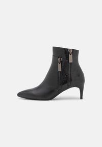 Bota negra de punta afilada hasta el tobillo con tacón delgado y dobles cremalleras verticales en el lado exterior sobre fondo blanco.